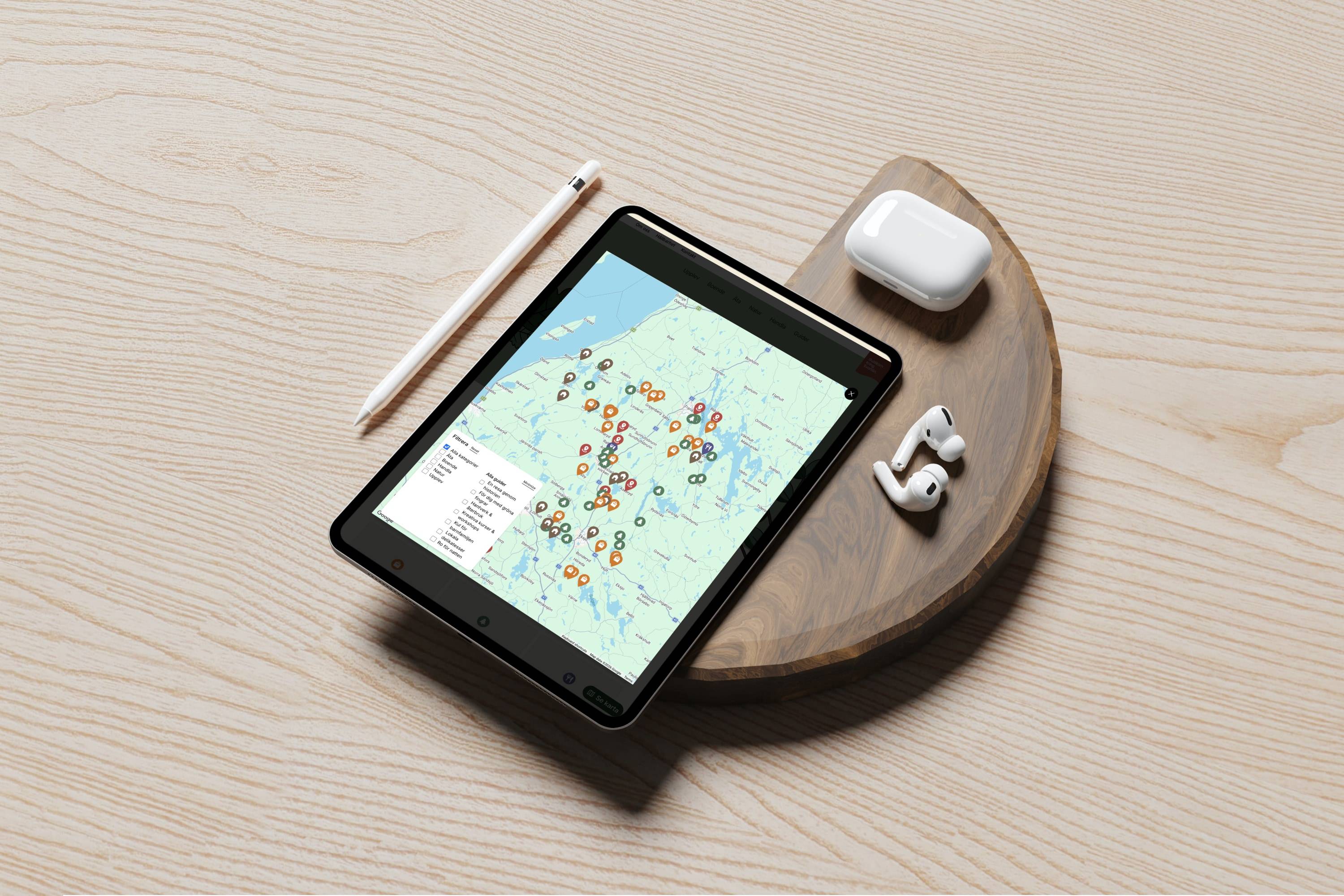The map open full screen on a tablet resting on a round wooden board, an Apple Pencil, an open AirPods case and a pair of AirPods beside it. Dozens of pins in category colours are spread across Smaland, with the filter panel in the lower left.